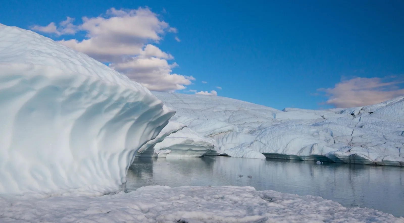 A curved ice arch beside a still meltwater pool under a bright blue sky