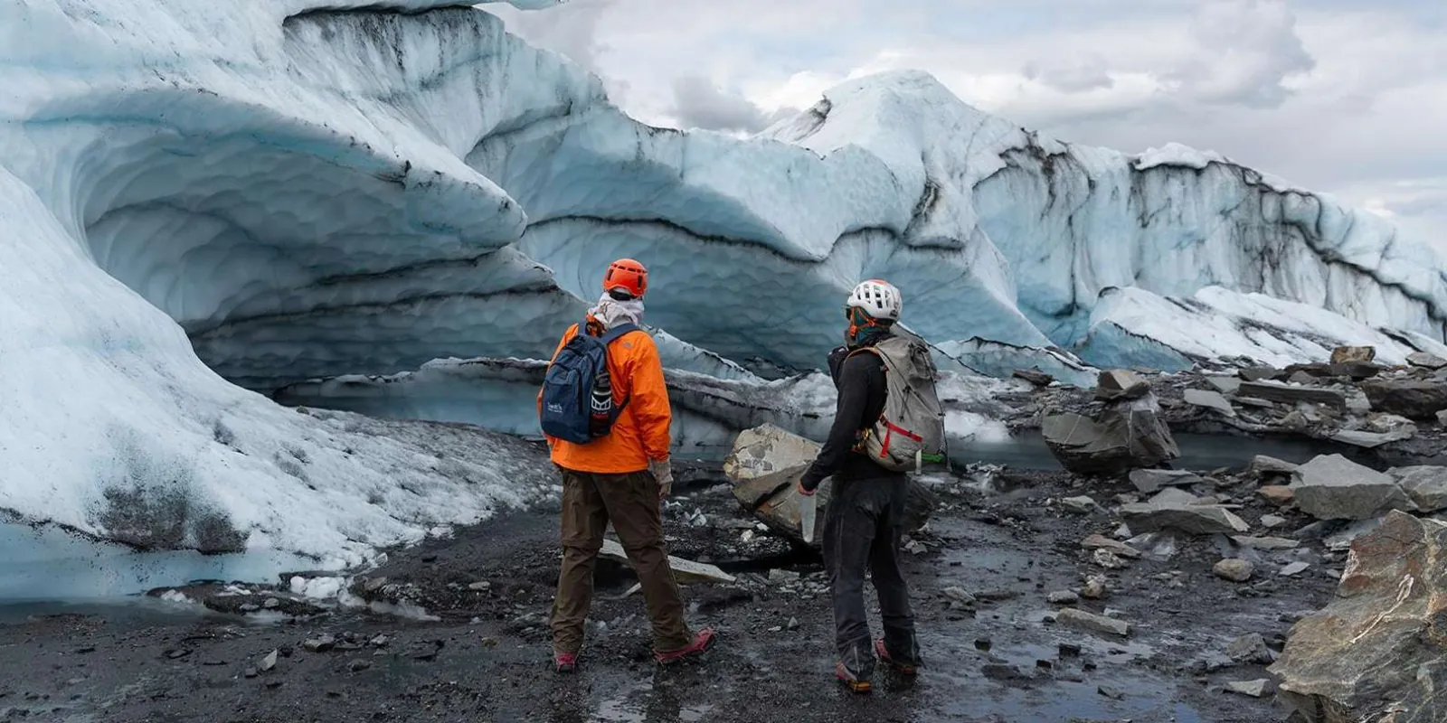 Two hikers in helmets beside a dark ice cave in the terminal moraine
