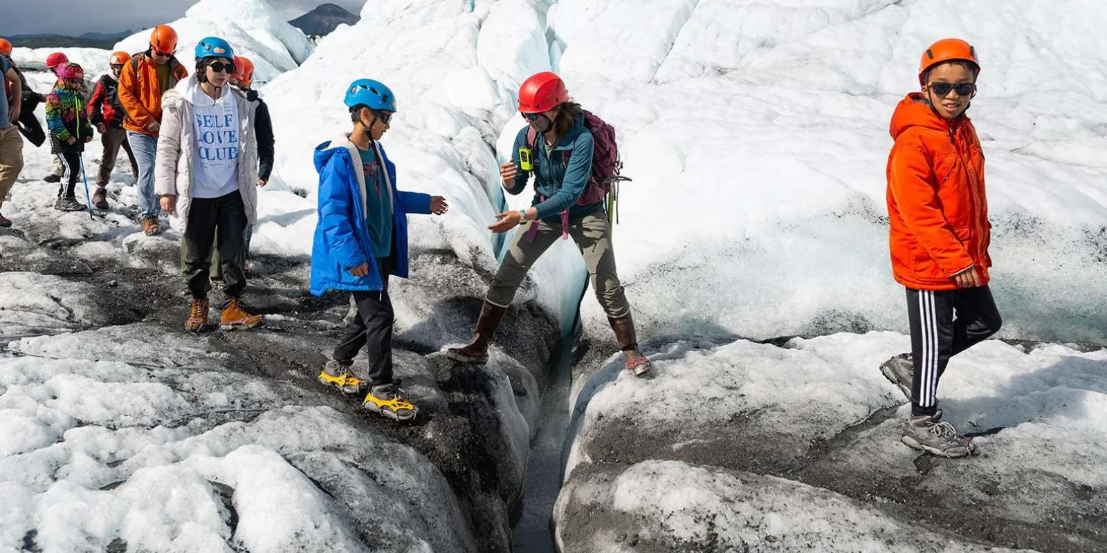 Children in helmets stepping across a shallow crevasse with their guide